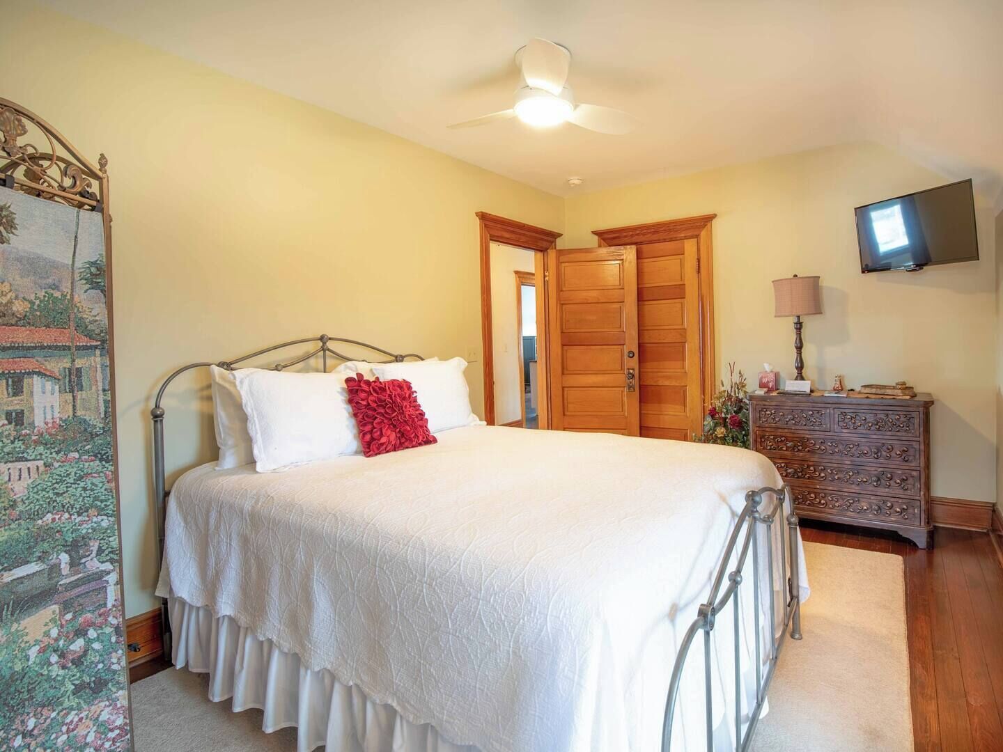 Une chambre à coucher comprenant un lit, un ventilateur de plafond, une armoire en bois, un téléviseur et une tapisserie décorative.