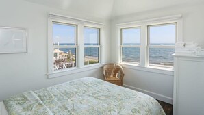 A bedroom with a bed, two windows with a view of the sea, a wicker chair, and a white dresser. - Truro Beachfront House For a Group! (Provincetown)