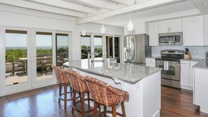 A modern kitchen with a large island, stainless steel appliances, and a view through the sliding glass doors. - Truro Beachfront House For a Group! (Provincetown)