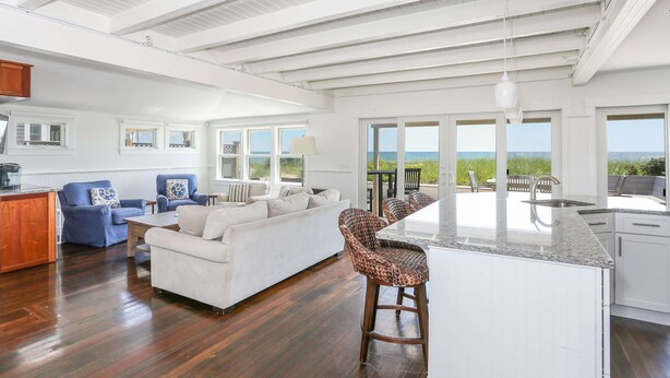 A spacious living area with a large white sofa, blue armchairs, and a kitchen with a bar area. - Truro Beachfront House For a Group! (Provincetown)