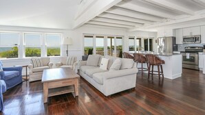 A spacious living area with a large sofa, a coffee table, and a kitchen area with modern appliances. - Truro Beachfront House For a Group! (Provincetown)