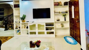 A modern living room with a flat-screen TV, white shelving, a glass coffee table, and a chandelier. - Cozy vacation home for family in Uptown Cagayan de Oro City Close to everything (Cagayan de oro city)