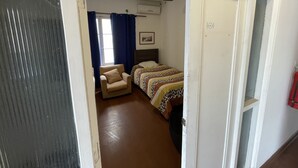 Un pasillo estrecho que conduce a un dormitorio con una cama, una silla y un ventanal con cortinas.