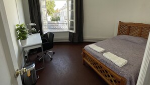 Una habitación pequeña con cama, escritorio y silla. Una ventana con cortinas da a una vista de edificios y un árbol.