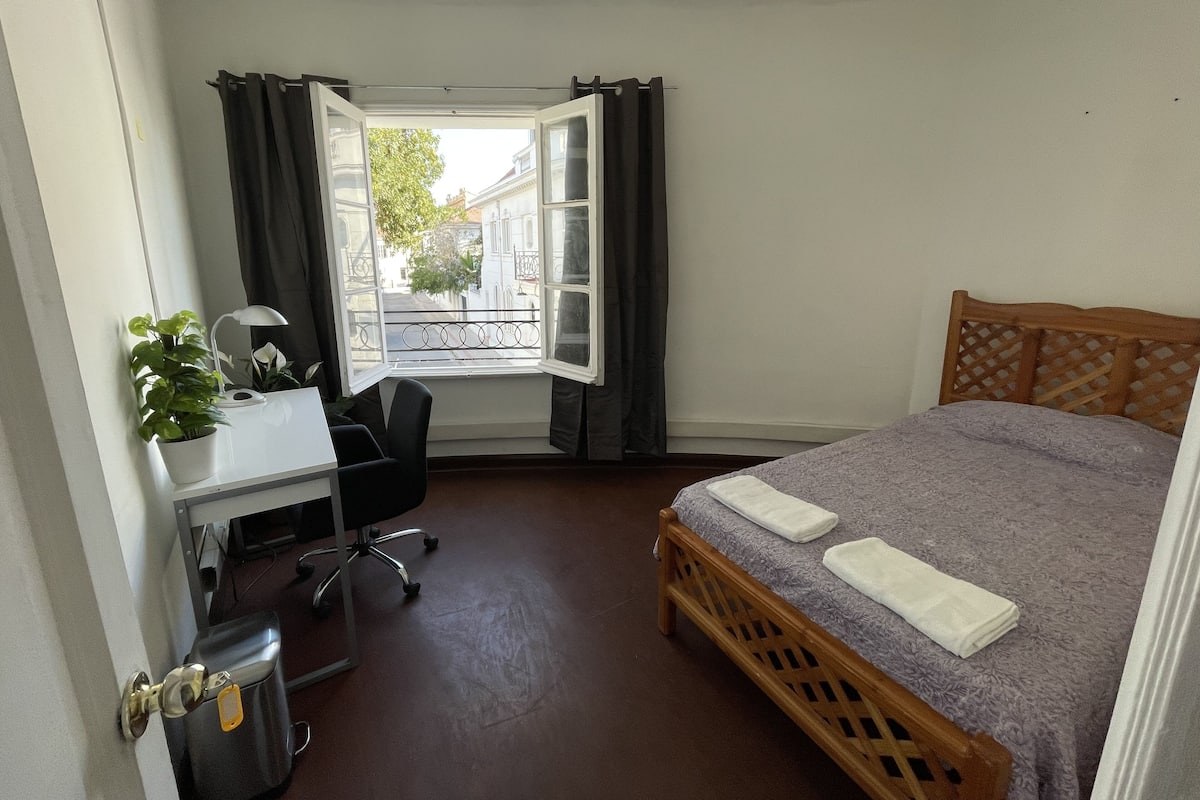 Une chambre à coucher de taille modeste, comprenant un lit, un bureau et une chaise. Une fenêtre, ornée de rideaux, offre une vue sur des immeubles et un arbre.