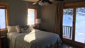 Ein Schlafzimmer mit Bett, Nachttisch, Deckenventilator, Fenster mit Jalousien, Lampe und Blick auf eine verschneite Landschaft durch eine Glastür.