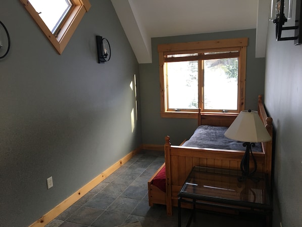 A bedroom with a sloped ceiling, a bed with a wooden headboard, a nightstand, a lamp, and a window with a view of trees.