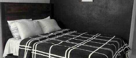 Una cama con una colcha a cuadros blancos y negros, almohadas blancas y un cabecero oscuro en una habitación con pared gris y un certificado o aviso enmarcado.