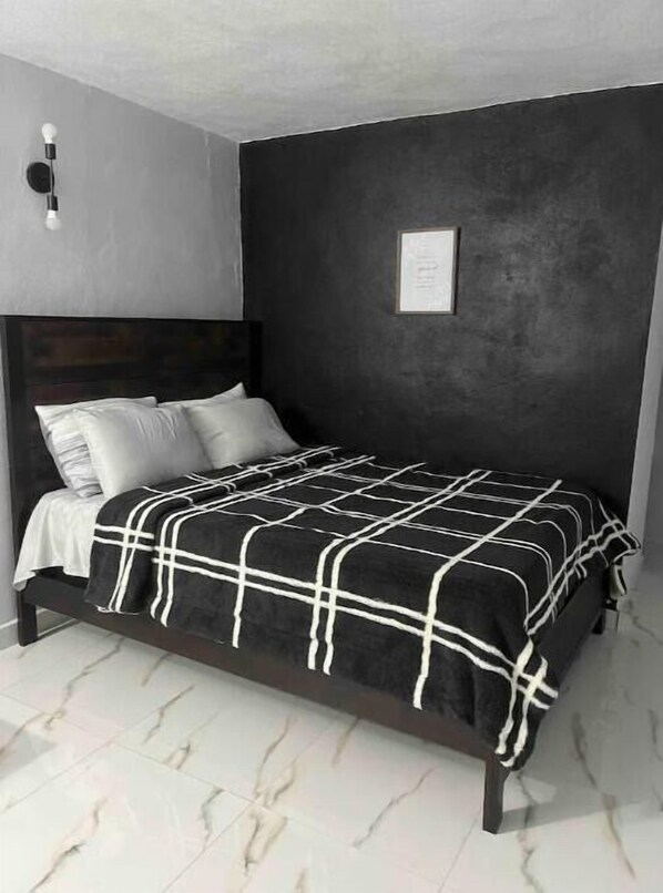 Un lit recouvert d’une couvre-lit à carreaux noirs et blancs, avec des oreillers blancs et une tête de lit sombre, dans une pièce dont un mur est gris et où est accroché un certificat ou un avis encadré.