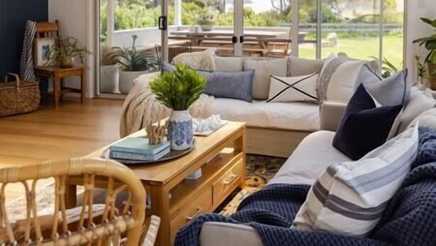 Una sala de estar acogedora con un sofá, una mesa de centro de madera y un sillón de mimbre. Se ve un jardín a través de amplias ventanas.