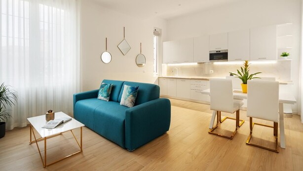 A modern living room with a teal sofa, a white coffee table, and a dining area with white chairs and a yellow plant. - Golden Teal apartment (BERGAMO)