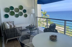 A balcony with a view of the ocean, furnished with wicker chairs, a white table, and decorative plates on the wall. - Beautiful Apartment in Sosua with Maid included (Sosua)