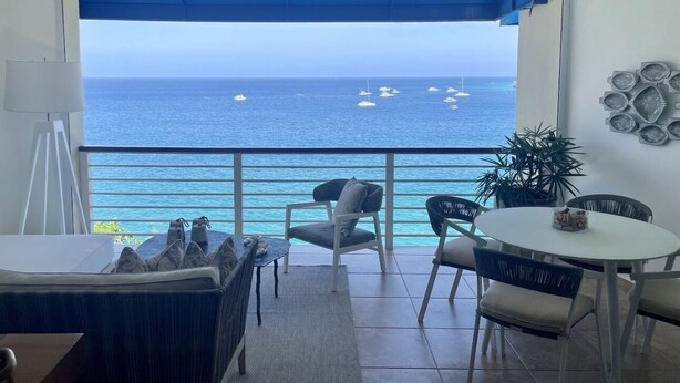 A balcony with a view of the ocean and boats, furnished with chairs and a table, and decorated with a plant and wall art. - Beautiful Apartment in Sosua with Maid included (Sosua)