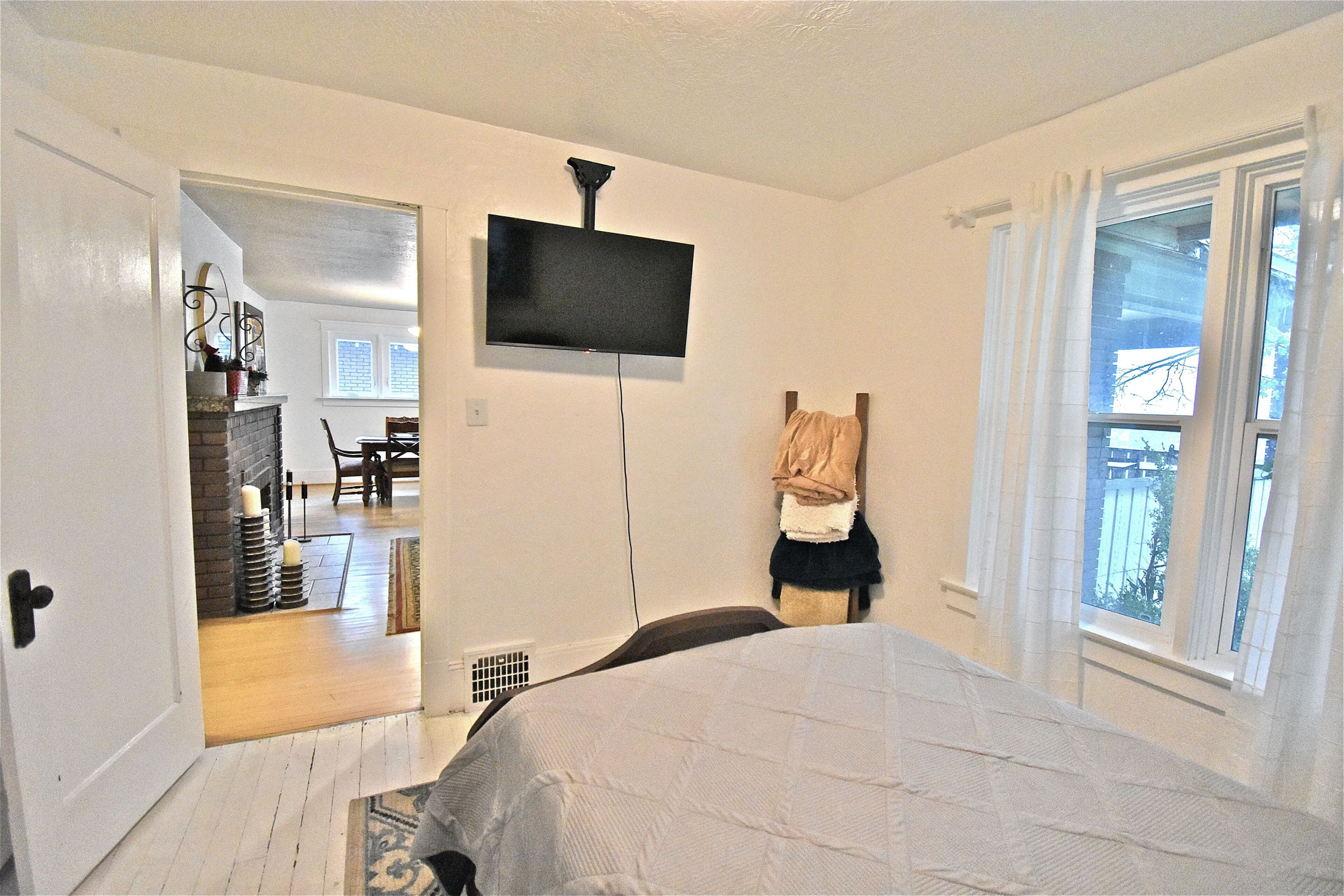 Un dormitorio con cama, televisor montado en la pared, ventana con cortinas y puerta que da a otra habitación.