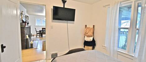 Un dormitorio con cama, televisor montado en la pared, ventana con cortinas y puerta que da a otra habitación.