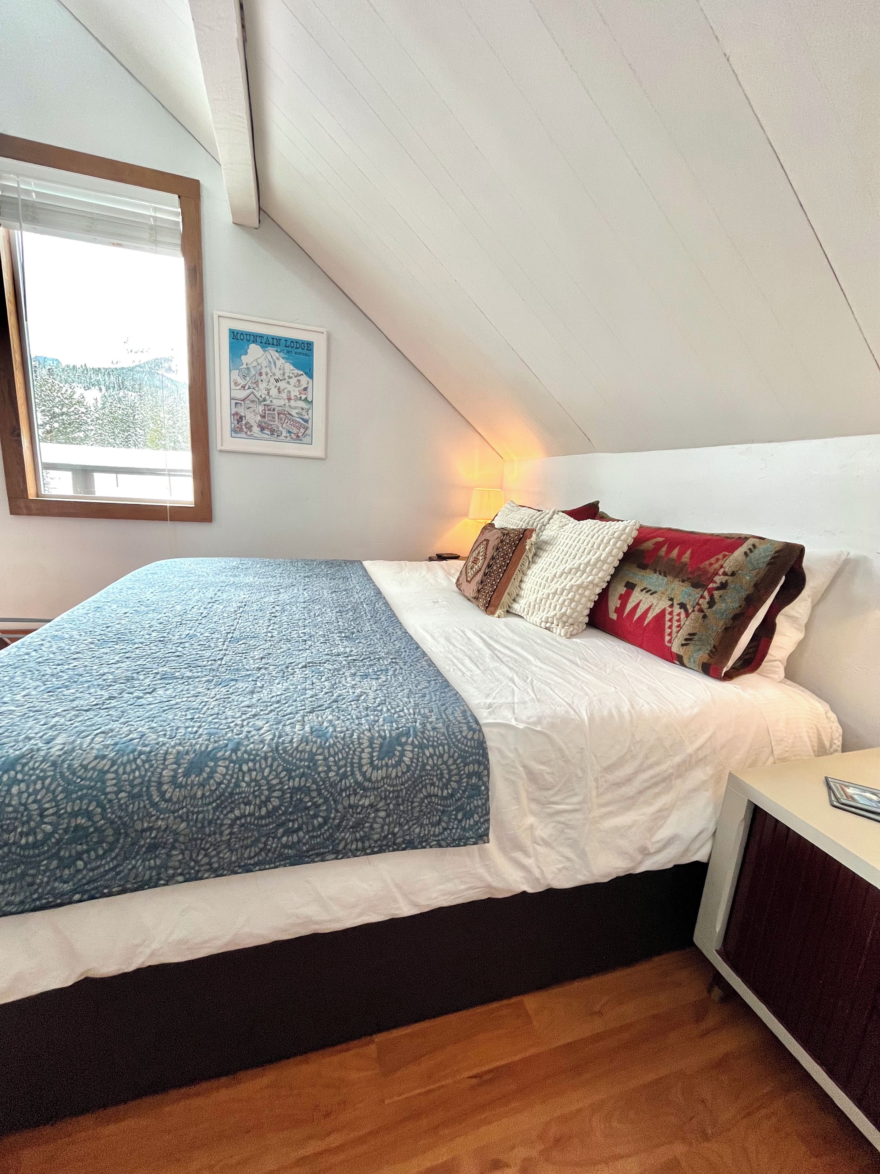 Une chambre à coucher avec un lit, une table de chevet, une lampe, un tableau encadré et une fenêtre avec des stores.