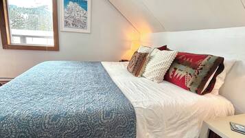 Une chambre à coucher avec un lit, une table de chevet, une lampe, un tableau encadré et une fenêtre avec des stores.