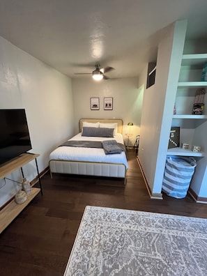 Een slaapkamer met een bed, een televisie, een plafondventilator, een nachtkastje en een wandplank met spullen.