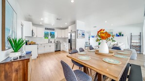A modern kitchen with white cabinets, a dining area with a wooden table and grey chairs, and a living area with a sofa and a coffee table. - Heated Pool+Hot Tub+Walk To Beach/Shops+Mini Golf! (Lauderdale-by-the-Sea)