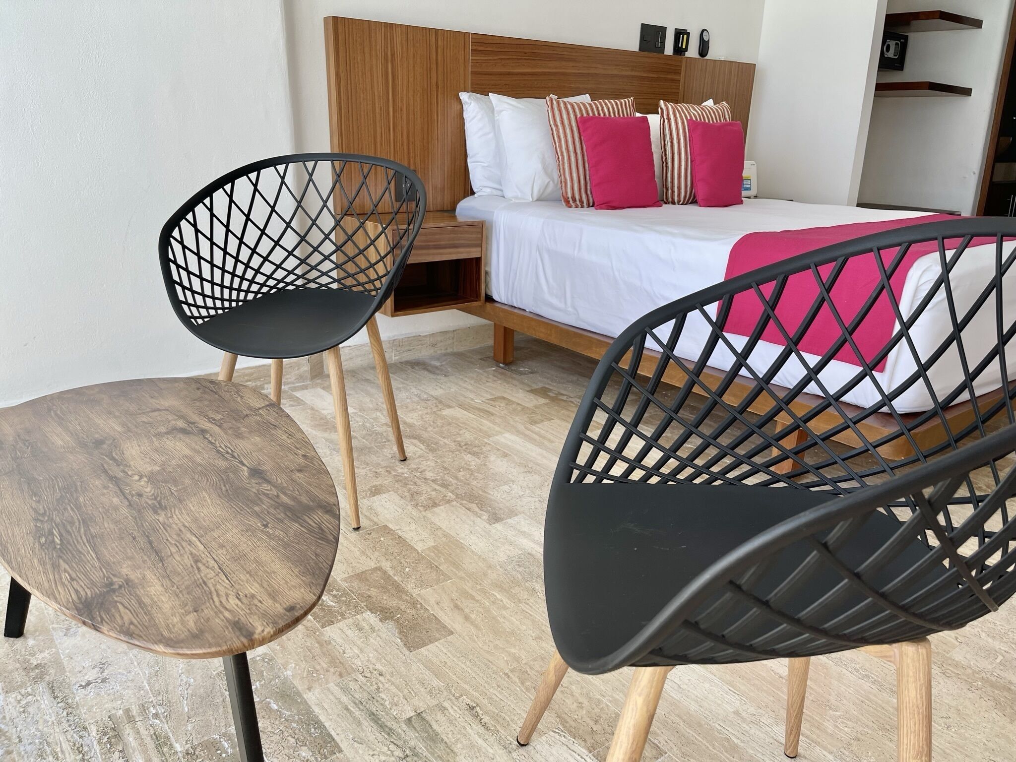 Une chambre à coucher avec une tête de lit en bois, un lit recouvert d’une literie blanche et rose, et deux chaises noires aux pieds en bois.