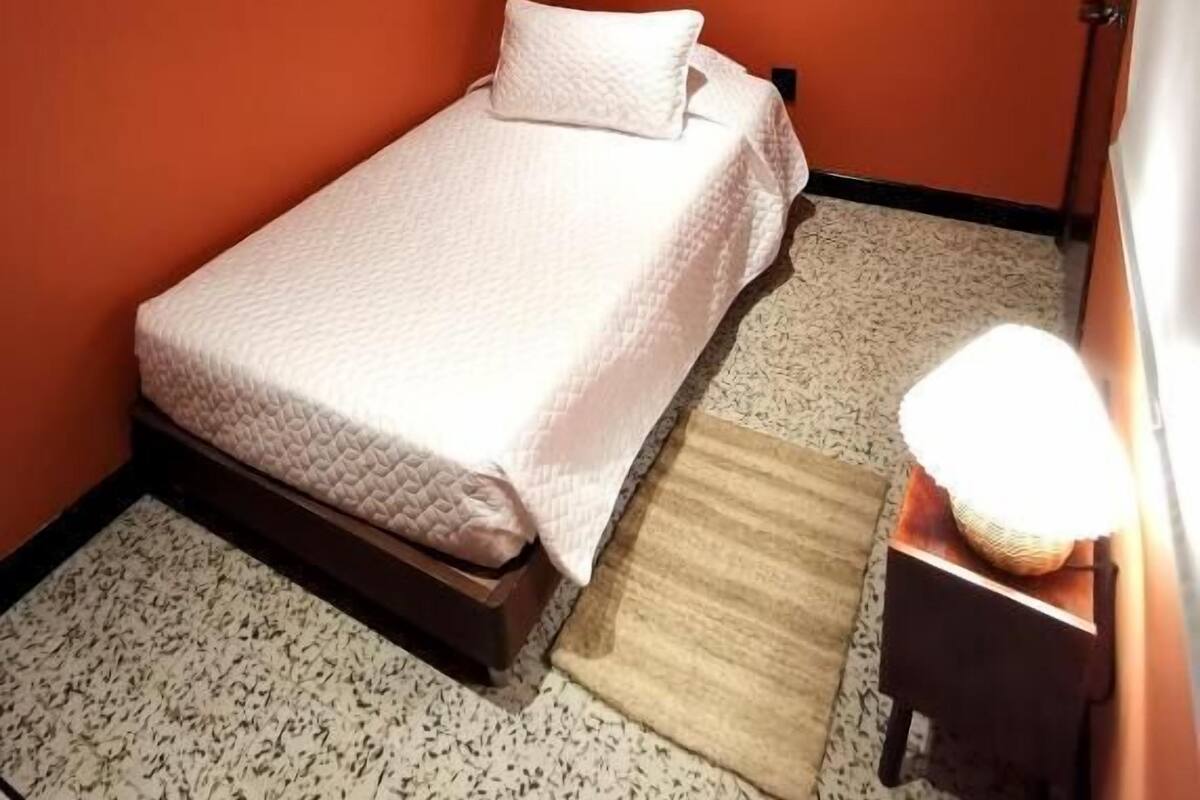 A small, single-bed room with a wooden bed frame, a bedside table with a lamp, and a patterned floor.