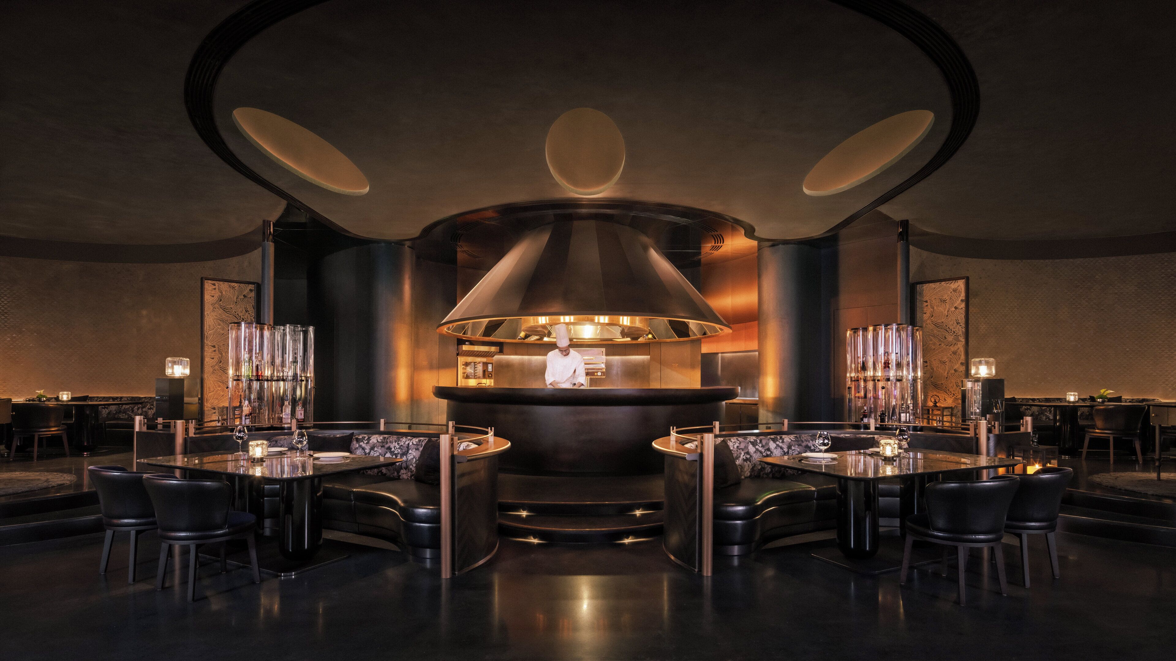 A modern restaurant interior with a central chef station, round tables, and black leather seating.