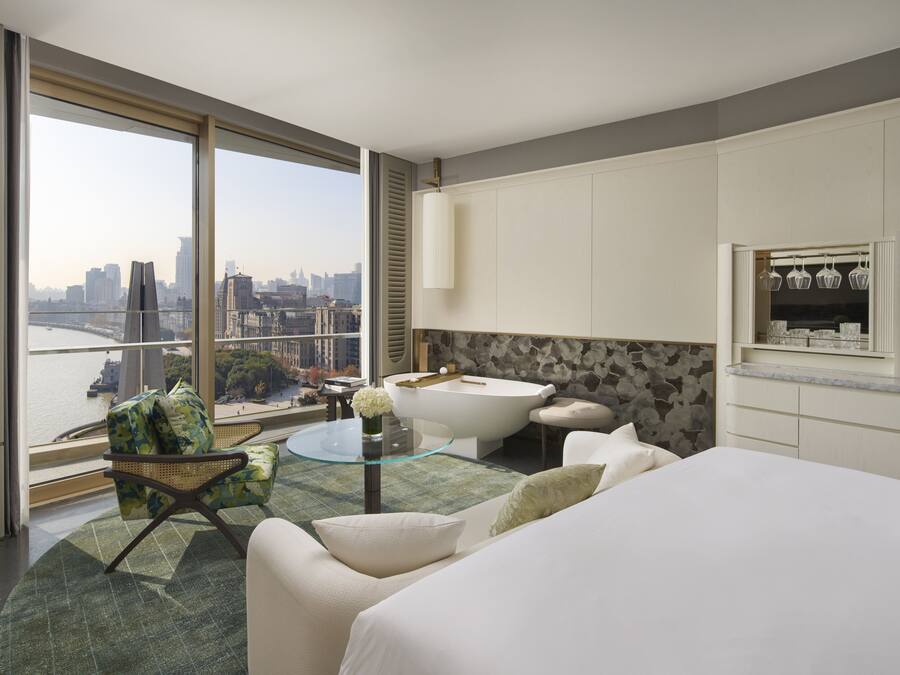 A modern hotel room with a large window offering a city view, a glass-top table, a sofa, a bathtub, and a kitchenette.