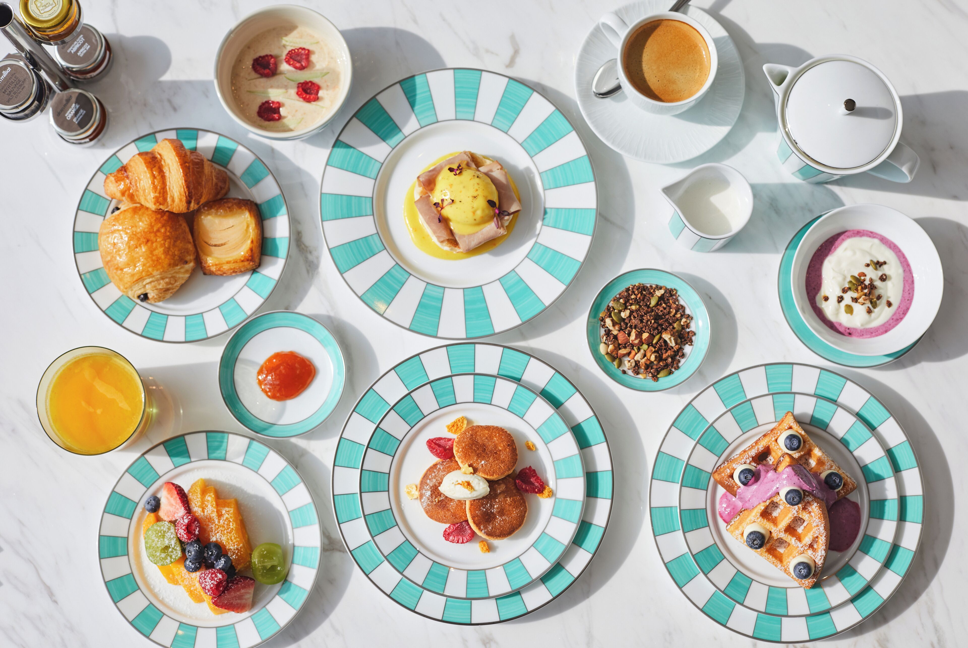 A breakfast setting with various dishes including pancakes, croissants, and fruit on plates, along with cups of coffee and a glass of orange juice.