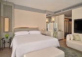A modern hotel room with a large bed, a sofa, a TV, and a bathroom with a mirror and vanity.