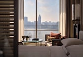 A hotel room with a large window offering a city skyline view, a round glass table with a plant, and a sofa with cushions.