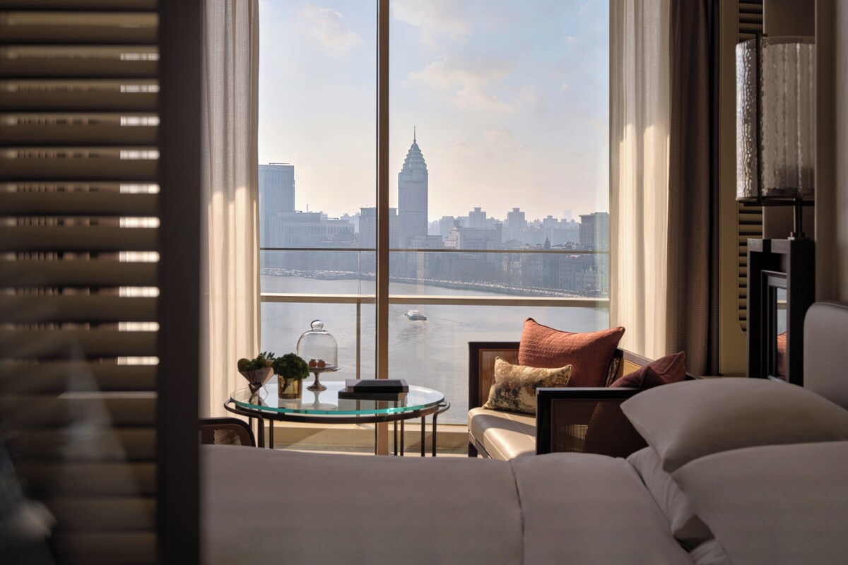 A hotel room with a large window offering a view of a city skyline and a river.
