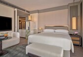 A hotel room with a large bed, a seating area with a glass table, a television, and a minibar.
