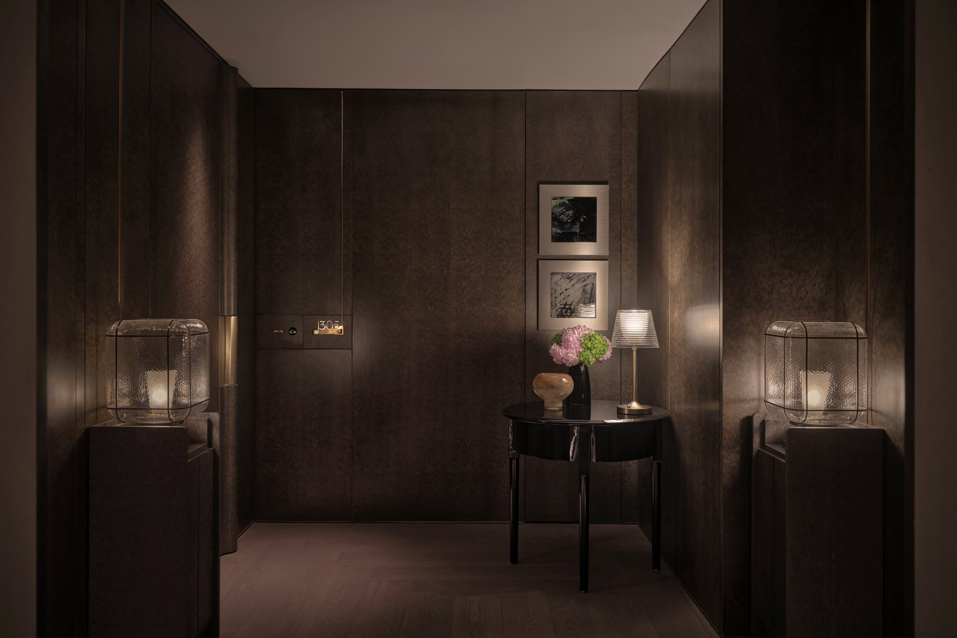 Un pasillo estrecho con paneles de madera oscura, una puerta con placa de número, una pequeña mesa con un jarrón y una lámpara, y dos lámparas de pared.