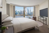 A modern hotel room with a large bed, a TV, a desk, and a view of a city skyline.
