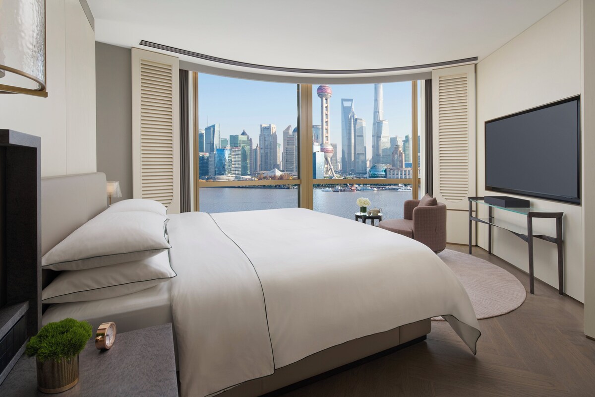 A modern hotel room with a large bed, a TV, a desk, and a view of a city skyline.