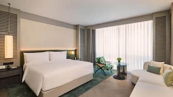 A modern hotel room with a large bed, a sitting area with a sofa and armchair, a small table with a plant, and large windows with blinds.
