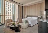 A modern hotel room with a large bed, a sitting area with a sofa and armchair, a glass coffee table, and a view of a cityscape through large windows.