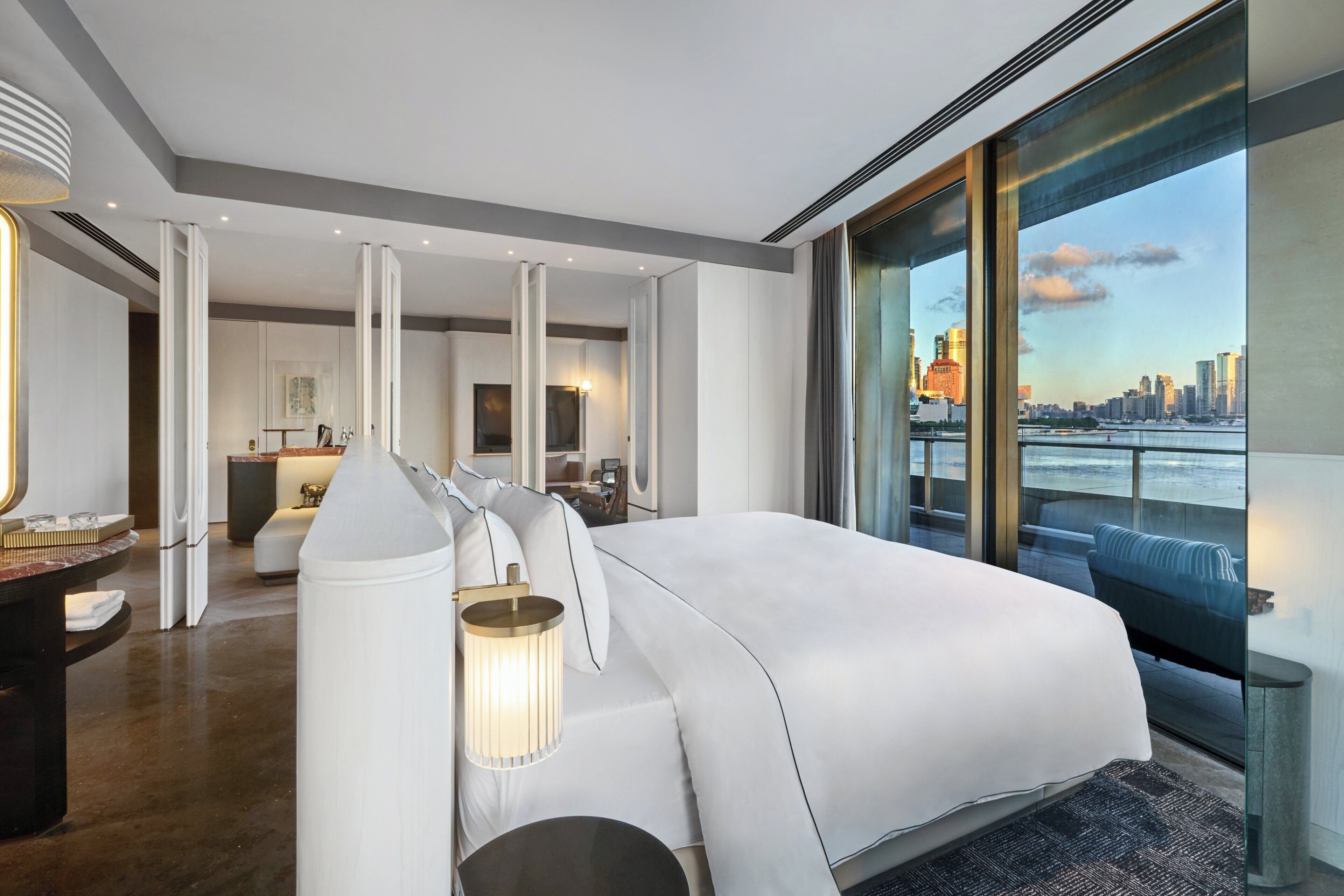 A modern hotel room with a large bed, a sitting area with a lamp, a view of the city through large windows, and a kitchenette in the background.