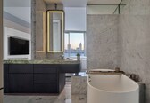 A modern bathroom with a large bathtub, a marble vanity, and a city view through the window.