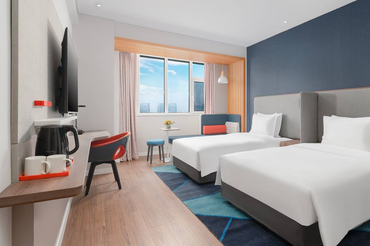 A hotel room with two beds, a desk with a coffee maker, a TV, and a window with curtains.