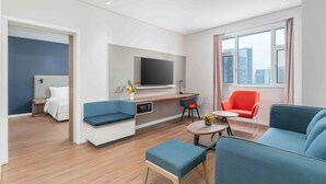 A modern hotel room with a sofa, a small table, a TV, and a window with a city view.