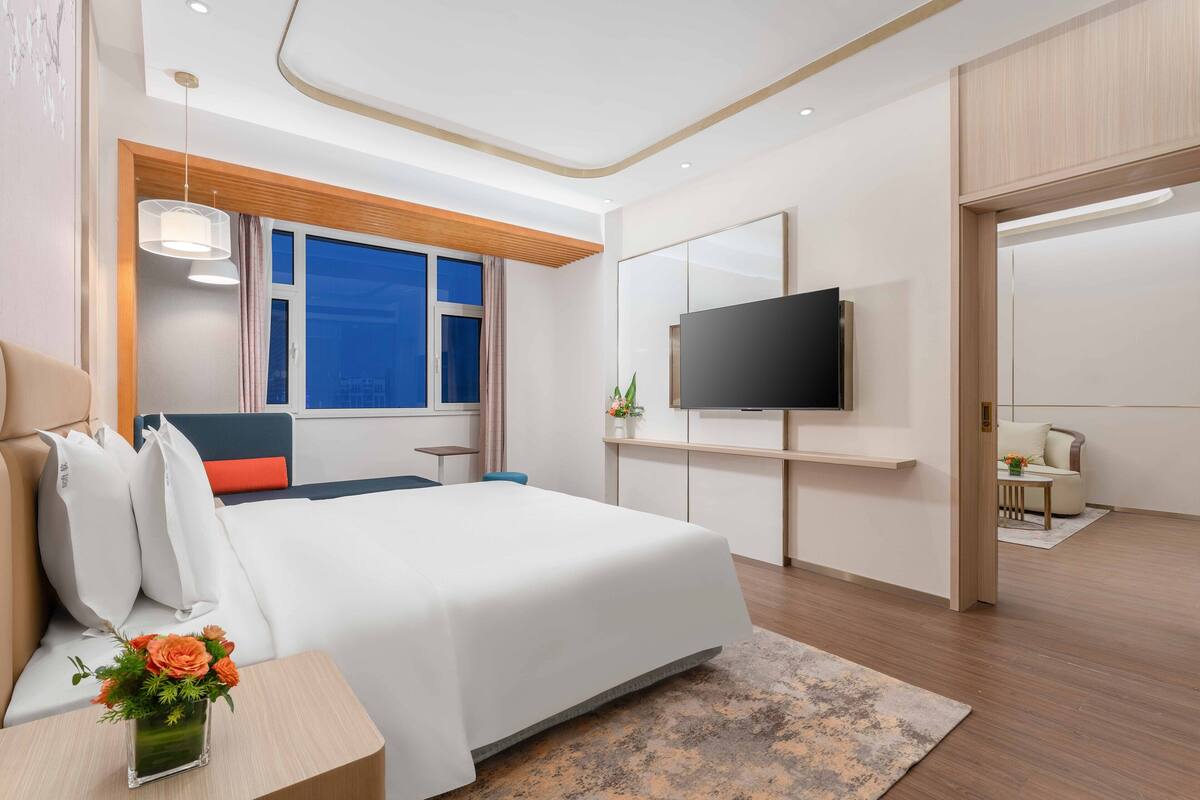 A modern hotel room with a large bed, a flat-screen TV mounted on the wall, a wooden ceiling, and a large window offering a view of the sky.