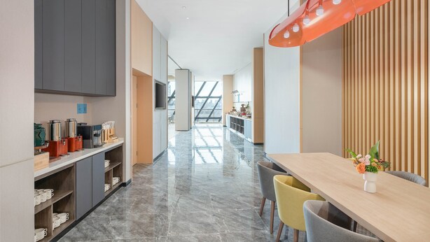 A modern hotel corridor with a long table and chairs, a coffee station, and a view to a balcony.