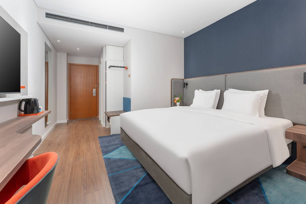 A hotel room with a bed, bedside tables, a desk with a chair, a TV, and a door.