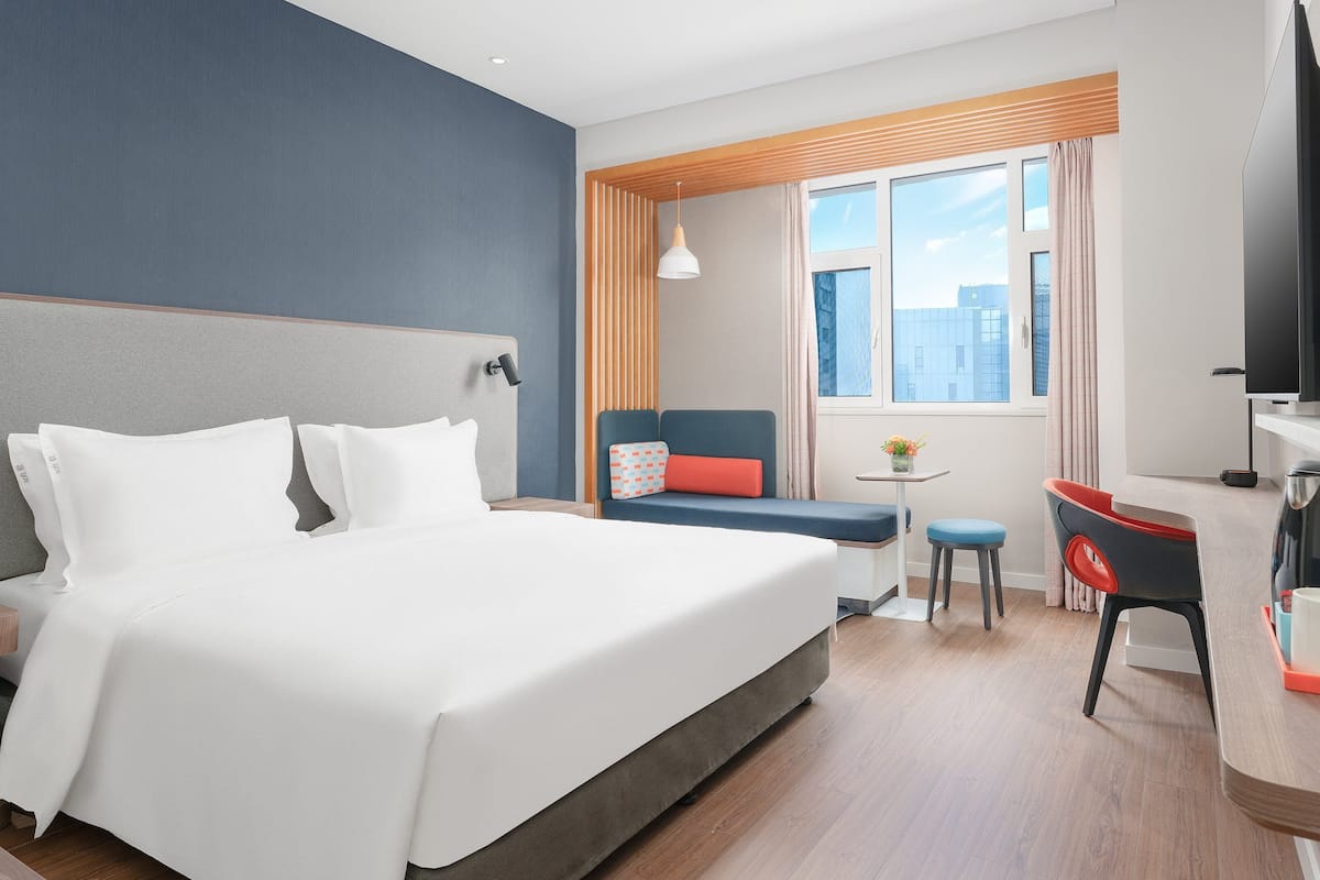 A hotel room with a large bed, a small table with a chair, a sofa, a TV, and a coffee machine.
