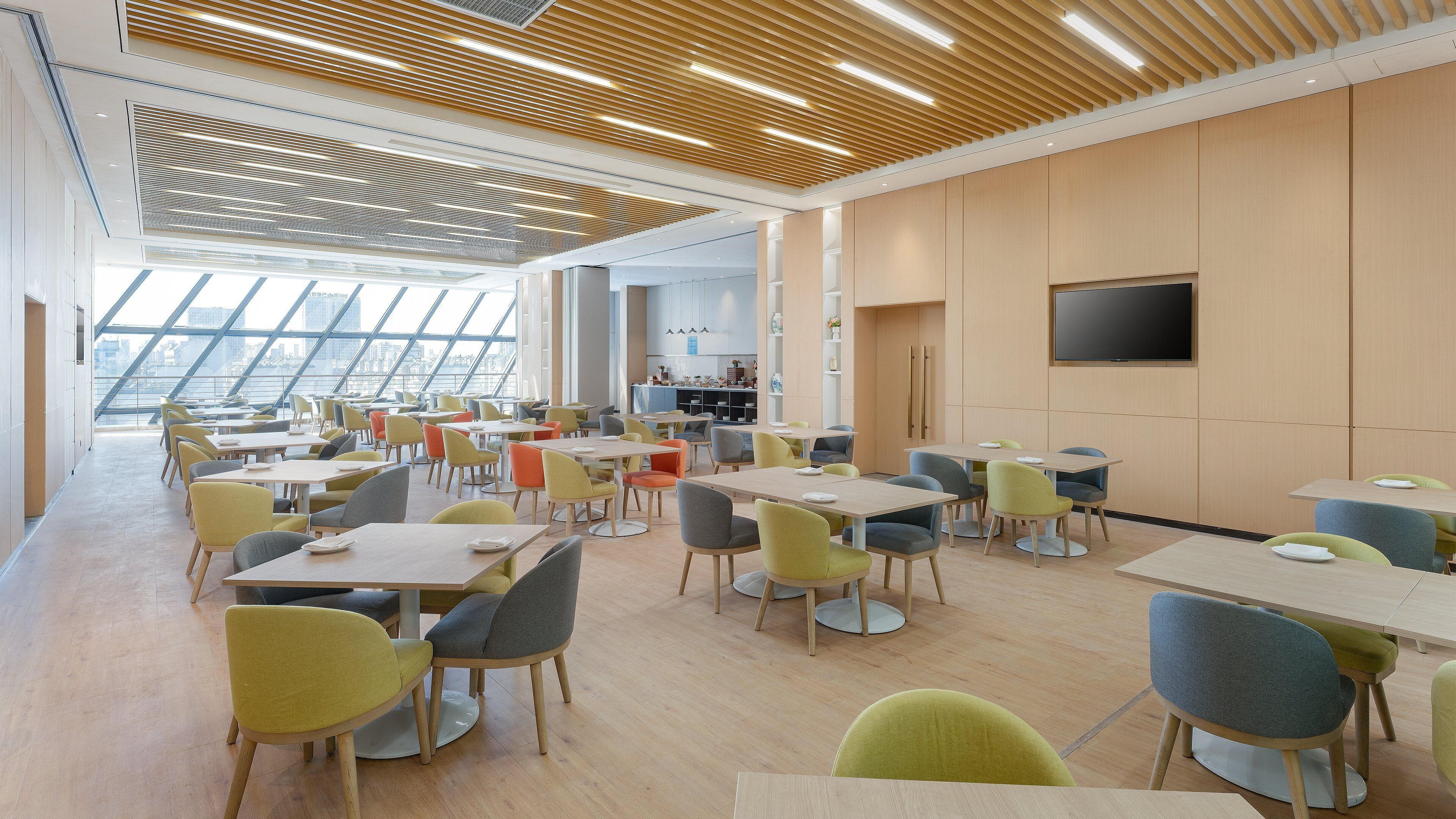 Un espace repas moderne avec plusieurs tables et chaises, une cuisine à l’arrière-plan et de grandes fenêtres offrant une vue sur la ville.