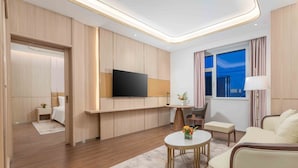A modern hotel room with a flat-screen TV mounted on the wall, a sofa, a small round table with a vase of flowers, and a window with curtains.