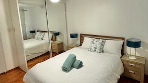 Una cama bien tendida con una colcha blanca y dos toallas enrolladas, un cabecero de madera, una mesita de noche con una lámpara y un ventilador de techo.