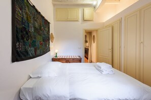Een slaapkamer met een groot bed, een wandtapijt aan de muur, een tafeltje met een lamp en een deur naar een andere kamer.
