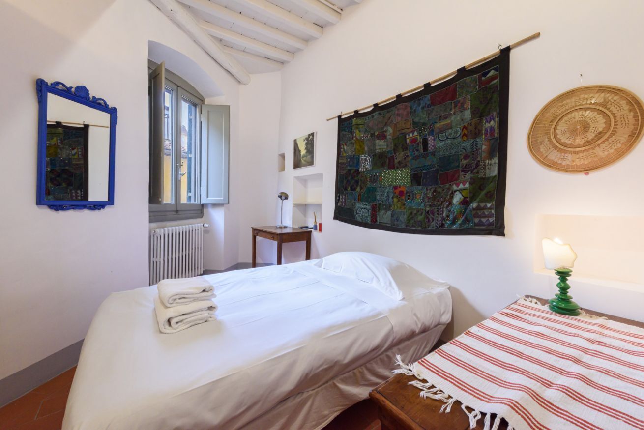 A bedroom with a bed, a small table with a lamp, a wall tapestry, a mirror, and a window.